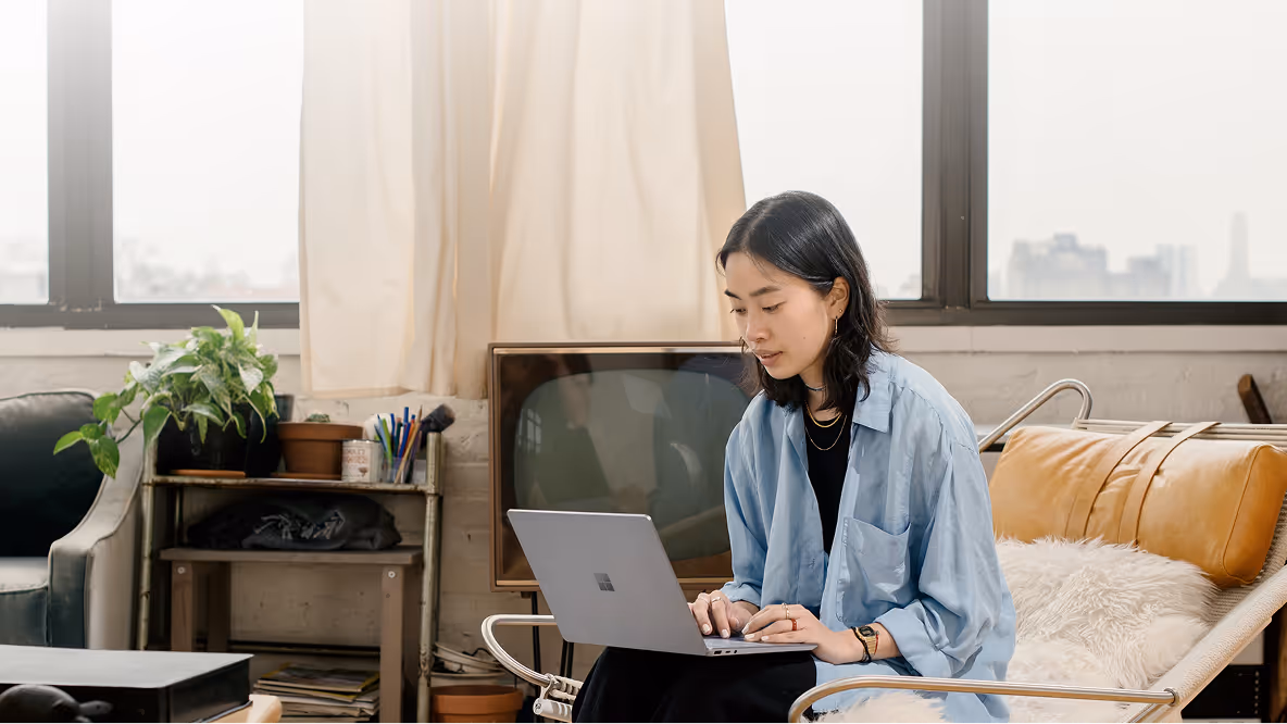 A profile view of a young woman with dark hair, wearing a thick, light blue knitted sweater, leaning over a table and writing in a notebook with a pen. In front of her is an open laptop displaying content, and to the left, a ring light on a stand with a smartphone holder suggests a video call or content creation setup. The background is a blurred, cozy indoor setting, possibly a living room.