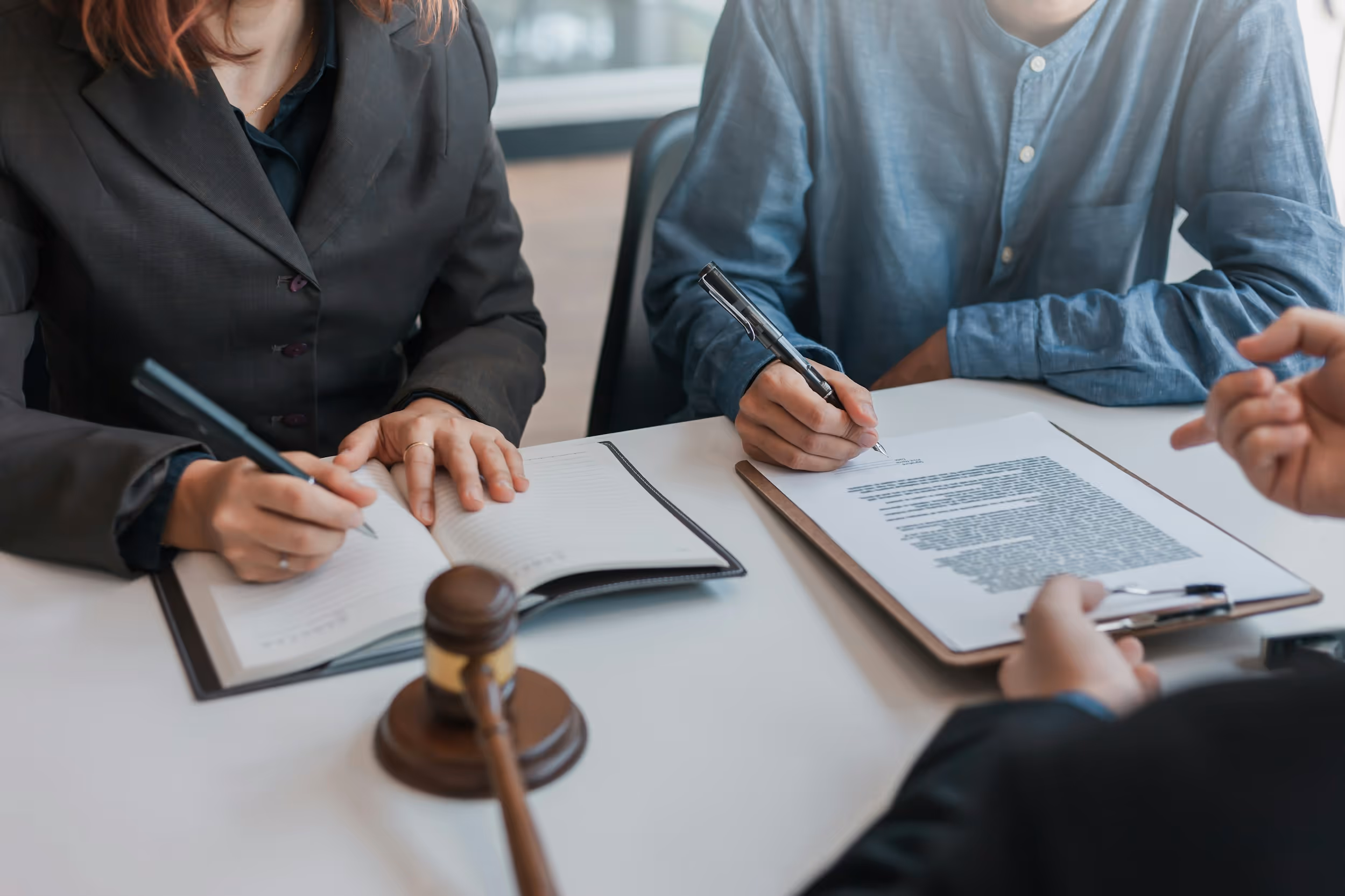 A medium close-up of three people gathered around a table, suggesting a legal or business meeting. A woman in a dark suit is writing in an open notebook on the left, and a person in a blue collared shirt is signing a legal document on a clipboard on the right. In the foreground, a third person's hand, wearing a dark suit jacket, points or gestures near a small wooden gavel, indicating a formal, perhaps contract-signing or mediation, setting.