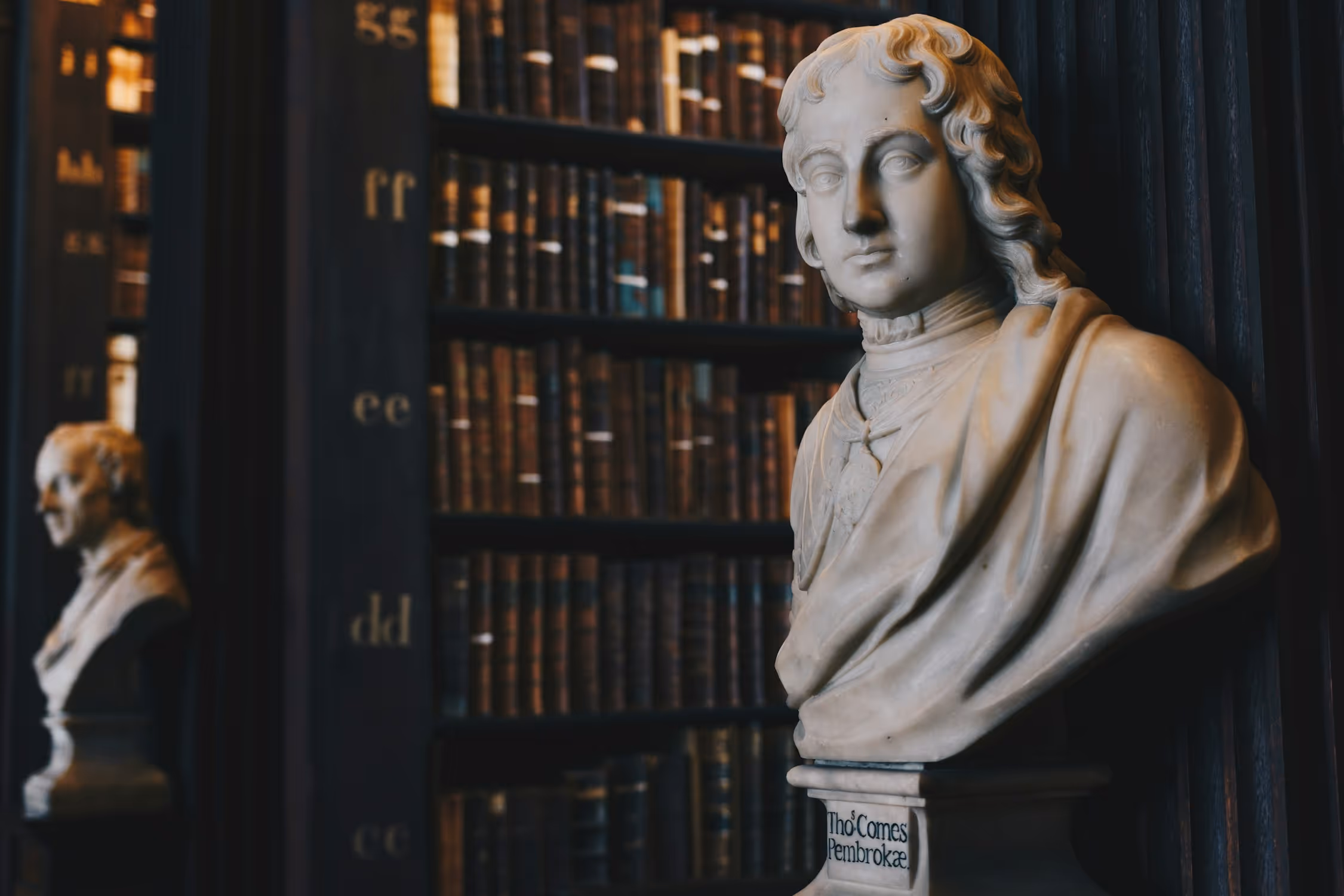 A detailed close-up of a light-colored marble bust of a classical or historical male figure, positioned in a dimly lit, traditional library. The bust is set against the backdrop of dark, floor-to-ceiling wooden bookshelves filled with old, leather-bound books. Another, less focused bust is partially visible in the reflection or background on the left.