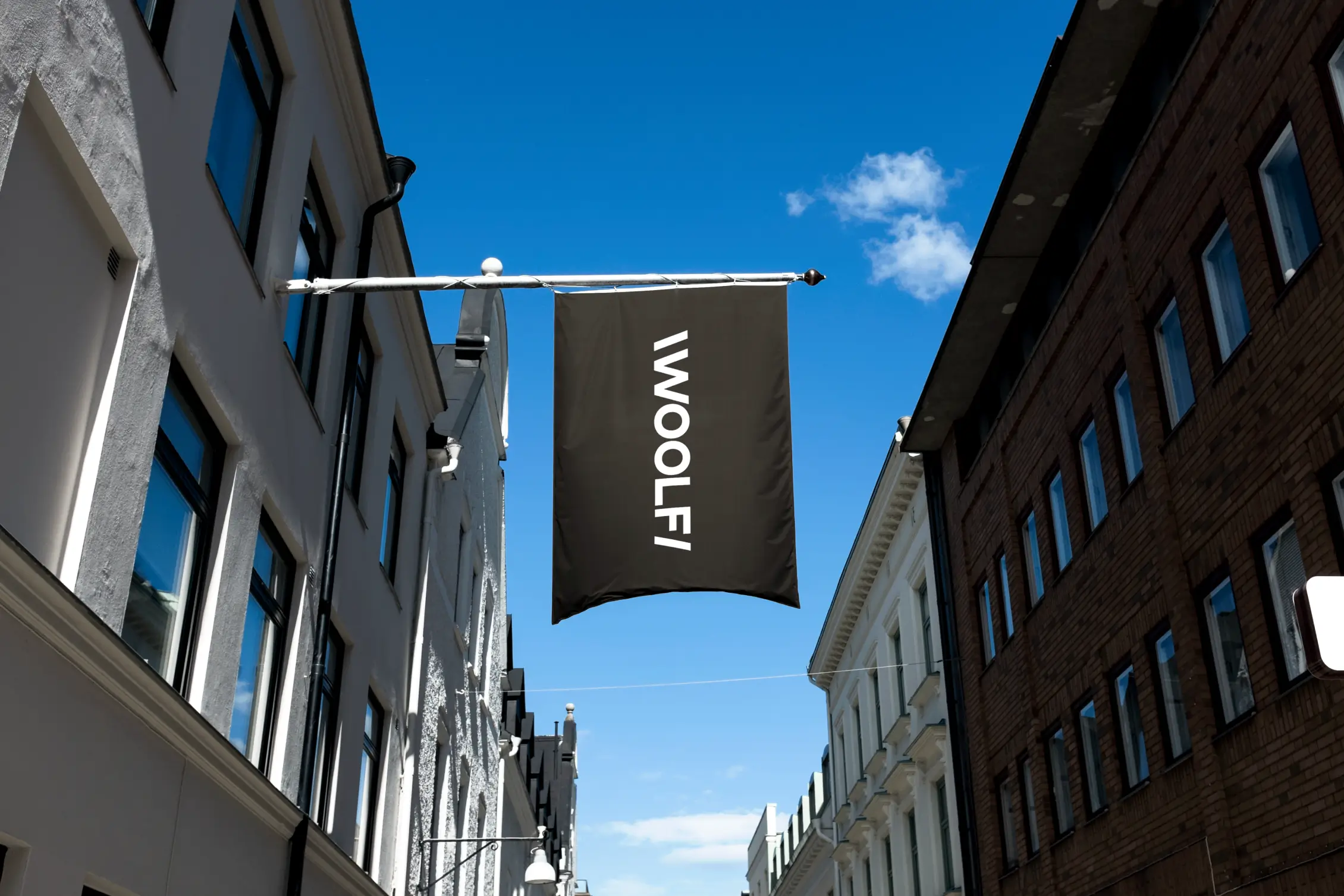 A vertical black banner flag displaying the word "WOOLF" in white capital letters, hanging from a pole attached to a building. The flag is centered in a narrow street between two rows of multi-story European-style buildings—one side white, the other side brown—under a bright blue sky with a few white clouds.