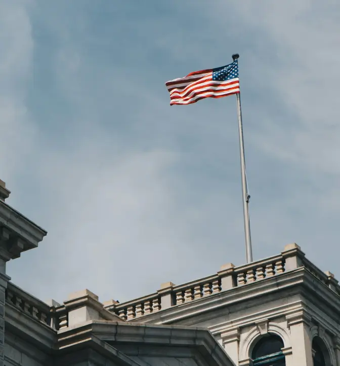 A low-angle shot of the American flag waving from a tall flagpole against a partly cloudy, light blue sky. The flagpole rises from the top of a stately, classical building facade, which features stone balustrades and detailed architectural elements.