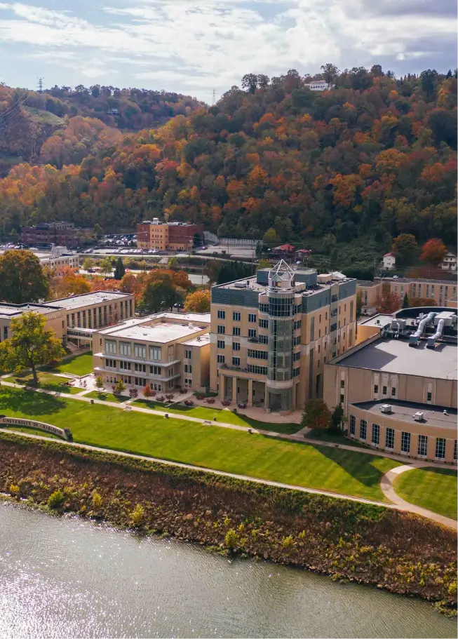An elevated view of a riverside campus or town area during the fall season. In the foreground, a grassy bank slopes down to a river. Dominating the middle ground are several large, tan and brick multi-story buildings, including a prominent modern structure with a curved, glass-heavy facade. The background features steep hillsides densely covered with trees showing vibrant autumn foliage in shades of orange, red, and yellow.