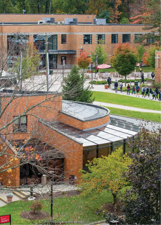 An aerial or elevated view of a university campus quad during autumn. The image is dominated by brick buildings and features a modern, curved structure in the foreground with a glass roof. In the background, a large academic building with a sign that reads "STUDENT CENTER" overlooks a walkway where a group of students is walking. The surrounding trees show fall foliage in shades of green, yellow, and red.